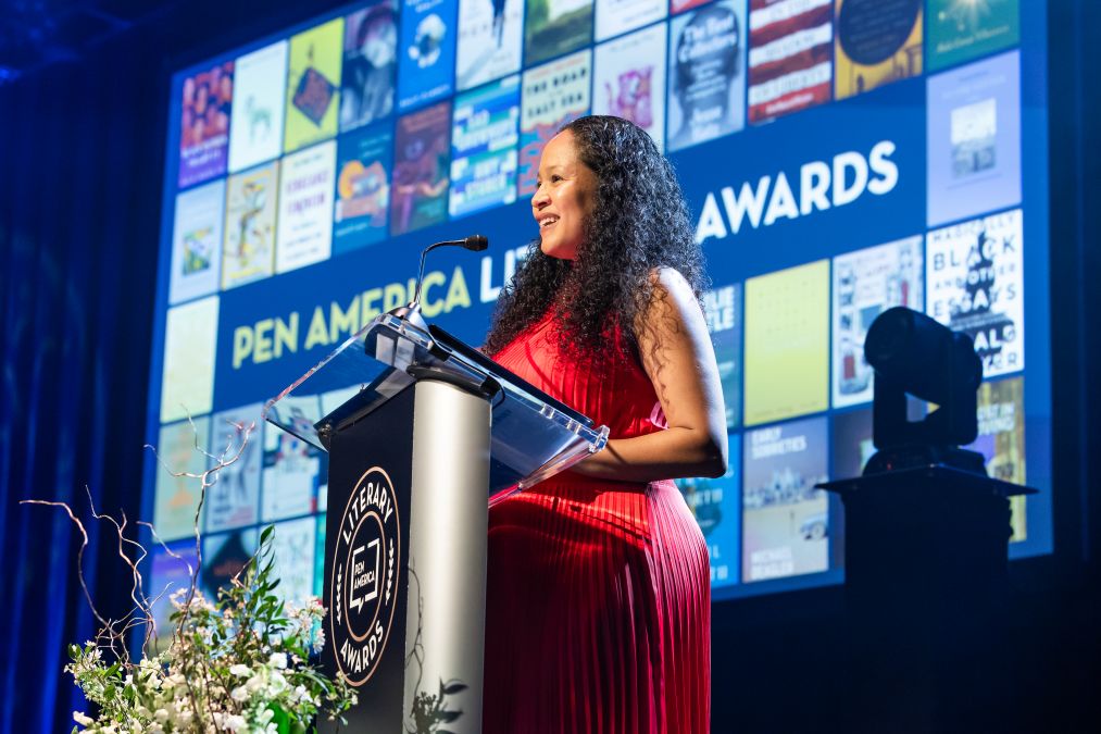 A woman in a red dress speaks at a podium during the PEN America Literary Awards ceremony, with a backdrop displaying the event name and images of book covers. Flowers are arranged in front of the podium.