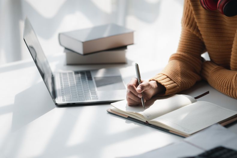 A person wearing a brown sweater writes in a notebook at a white desk with an open laptop and stacked books nearby. Sunlight casts shadows across the workspace.