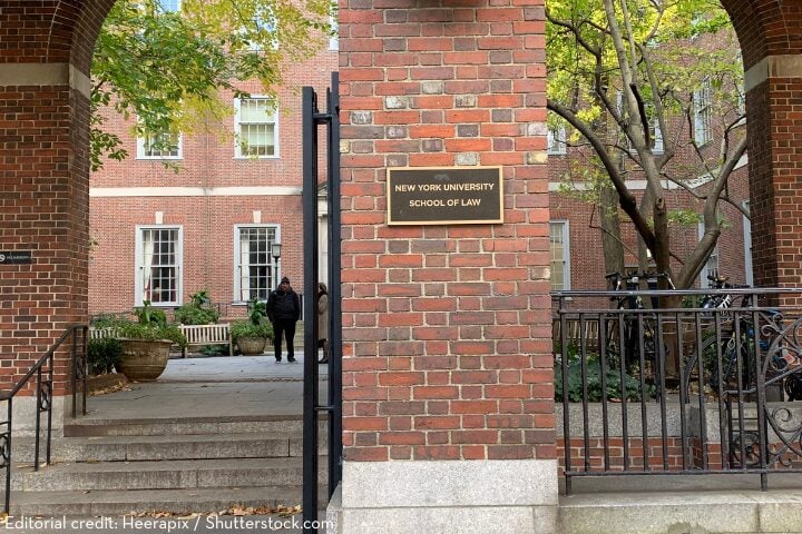 A brick archway with a sign reading New York University School of Law opens to a courtyard, where two people stand near potted plants and benches. Bicycles are parked to the right, and trees provide partial shade.