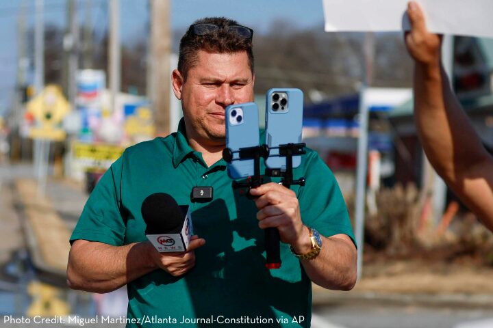 A man in a green shirt holds a microphone and a smartphone rig with two phones, recording outdoors. He appears focused, with a blurred street scene in the background.