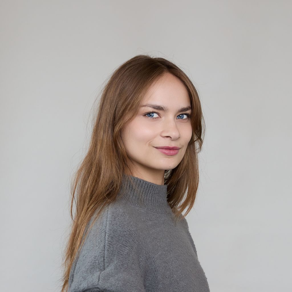 A young woman with long light brown hair and blue eyes, wearing a gray sweater, stands in front of a plain light background and smiles softly at the camera.