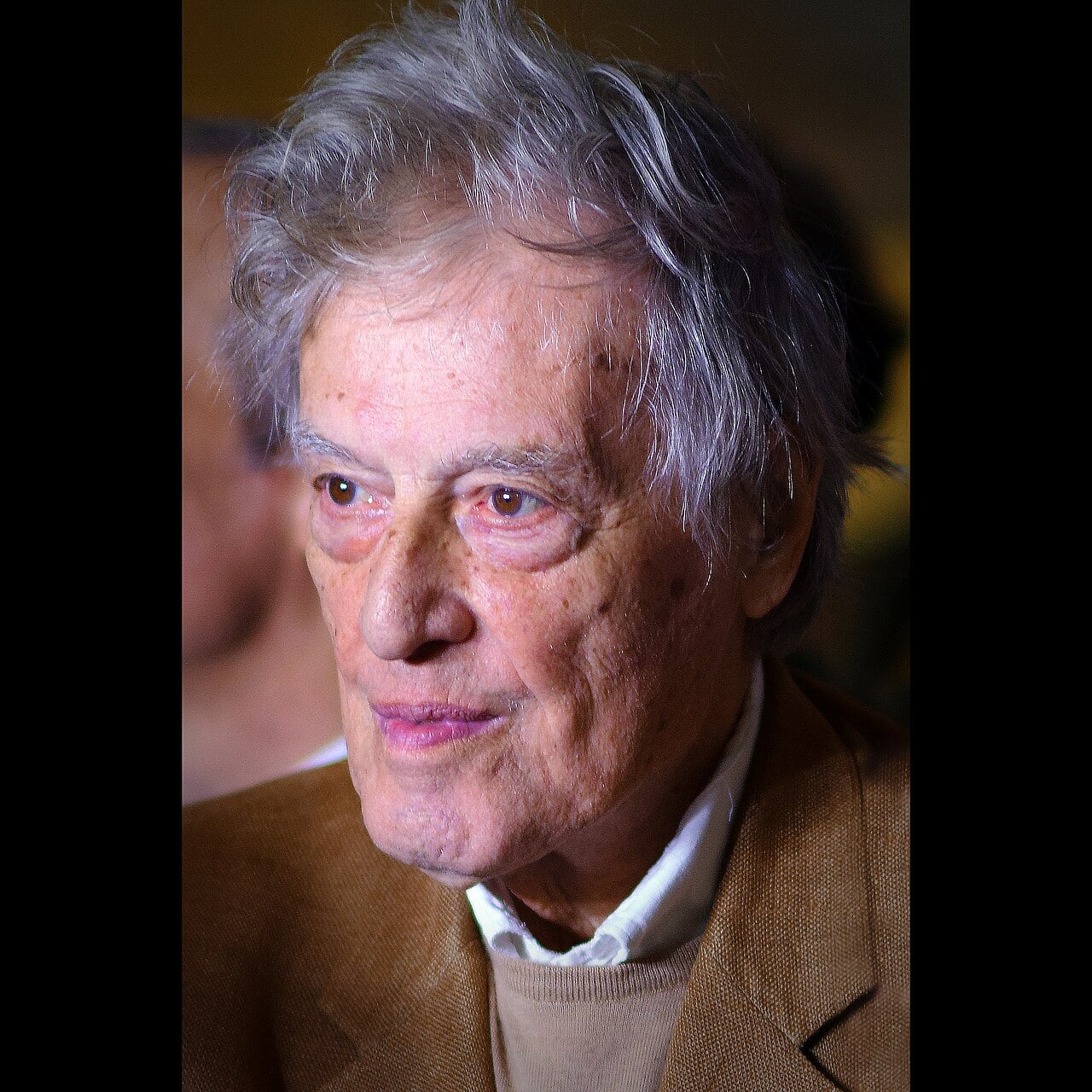 An elderly man with gray, tousled hair and a light brown jacket looks slightly to the side. The lighting highlights his face, showing a thoughtful expression and slight smile. The background is blurred.