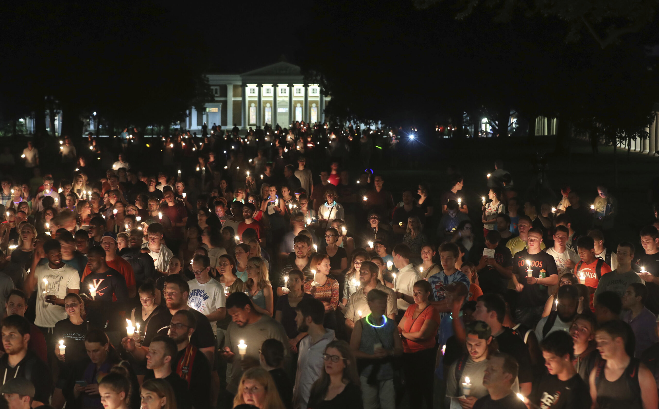 A large crowd gathers outdoors at night, holding lit candles in a vigil. The background features a well-lit building with columns, and the atmosphere appears solemn and reflective.