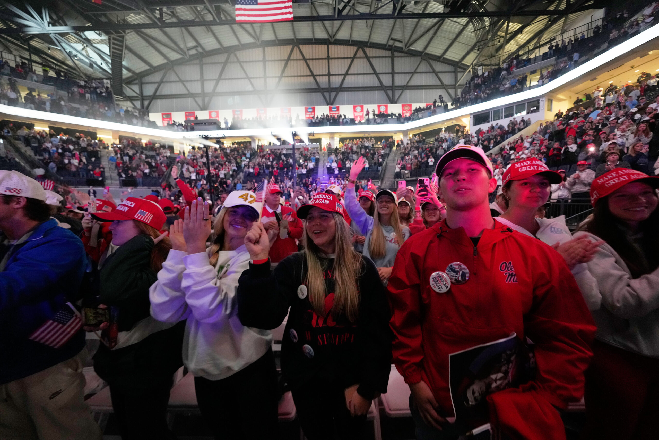A large crowd at an indoor rally, many wearing red Make America Great Again hats and holding flags, with bright lights and a full audience in the stadium seats in the background.