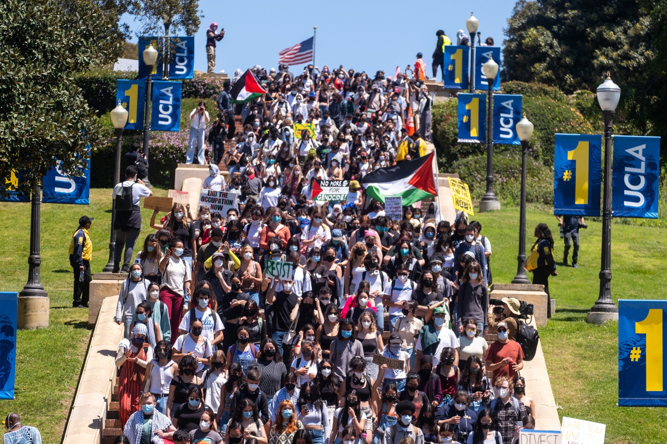 A large crowd of protesters holding signs and flags march down a staircase at UCLA, surrounded by blue banners with “UCLA” and “#1” on them; some people wear masks and an American flag waves in the background.