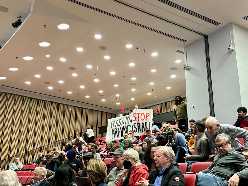 Audience seated in a lecture hall, with two people at the back holding a sign that reads Raskin STOP ARMING ISRAEL. The mood appears attentive, and some people are looking toward the sign.