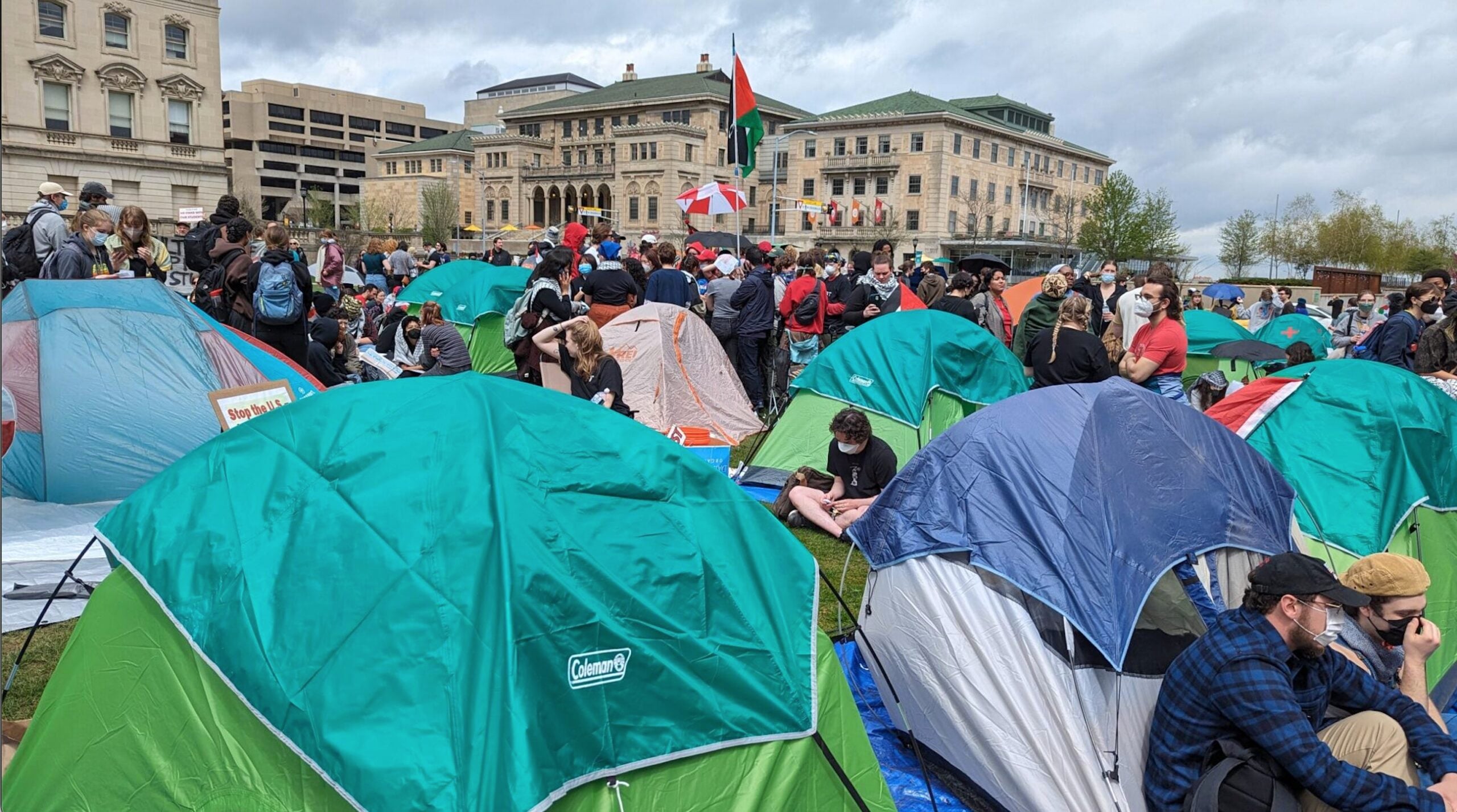A large group of people gather among numerous tents on a grassy area in front of university buildings, with some holding flags and signs; the sky is cloudy.
