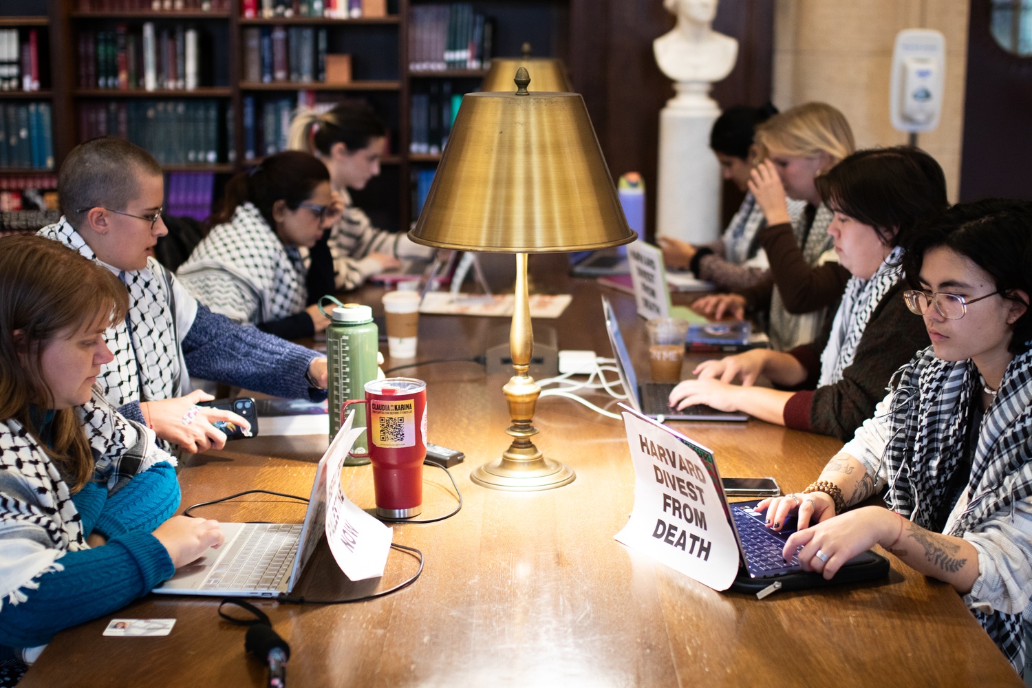 A group of people wearing black-and-white patterned scarves work on laptops around a library table with a lamp. Signs on the table read “HARVARD DIVEST FROM DEATH.” Bookshelves are visible in the background.