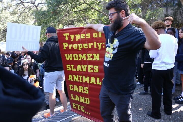 A man at a protest holds a red and yellow sign listing Types of Property: Women, Slaves, Animals, Cars, Land, etc. Other people and trees are visible in the background.