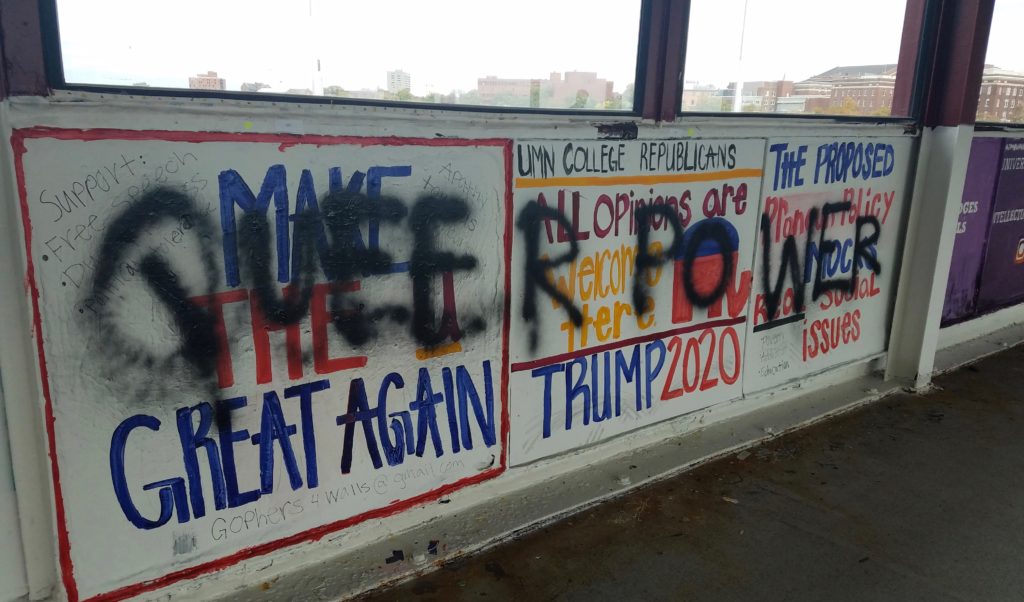 A wall mural featuring Make America Great Again and pro-Trump slogans is defaced with black spray paint obscuring parts of the text. Messages about campus Republicans and political issues are partially visible.