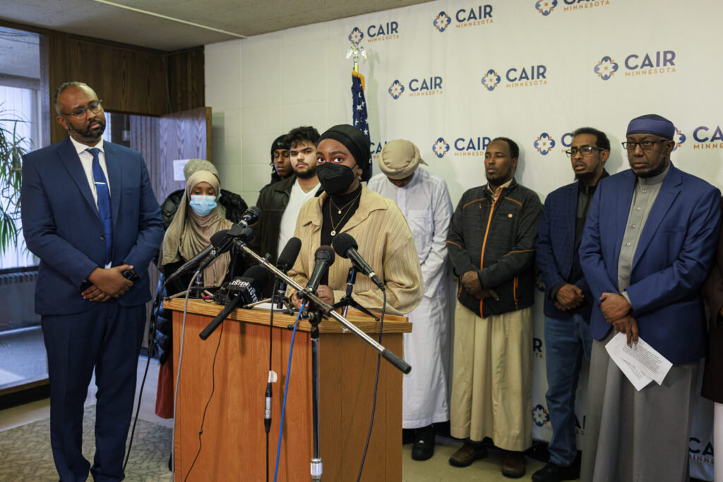 A woman in a headscarf speaks at a podium with microphones, surrounded by a group of people in traditional attire at a CAIR Minnesota press conference. An American flag is visible in the background.