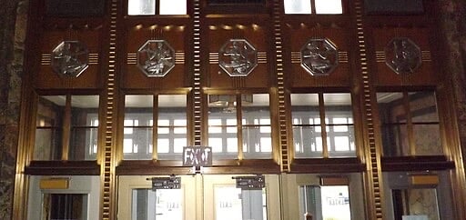 Art Deco-style interior doors with decorative metallic reliefs above, featuring geometric patterns and figures. The doors have glass panels and an illuminated EXIT sign above the center doorway.