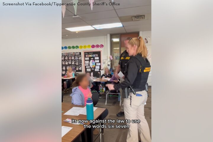 A sheriffs deputy stands in a classroom speaking to students. One student sits at a desk; faces are blurred. Classroom walls have colorful decorations. Text at the bottom reads, its now against the law to use the words six seven.