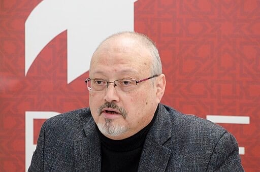 A middle-aged man with glasses, a mustache, and a goatee is wearing a black turtleneck and gray blazer, speaking in front of a red background with white geometric patterns and shapes.