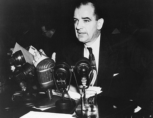 A man in a suit speaks behind several microphones at a table, gesturing with one hand. Papers and a glass of water are in front of him. The setting appears formal, possibly a hearing or press conference.