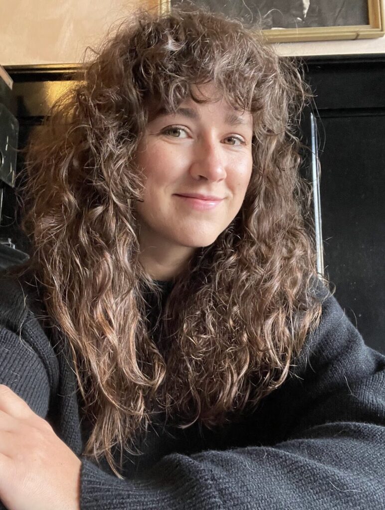 A person with long, curly brown hair and wearing a black sweater smiles at the camera while sitting indoors against a dark-colored wall.