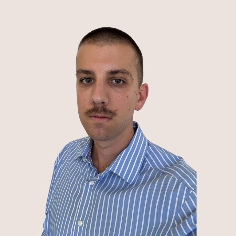 A man with short brown hair and a mustache, wearing a blue and white striped shirt, is looking at the camera against a plain light background.