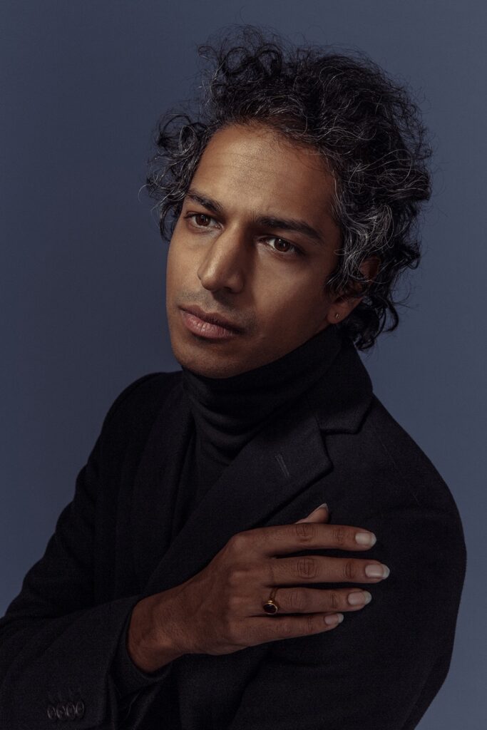A person with wavy, graying hair wearing a black turtleneck and blazer poses against a dark blue background, looking thoughtfully into the distance with one hand resting on their shoulder.