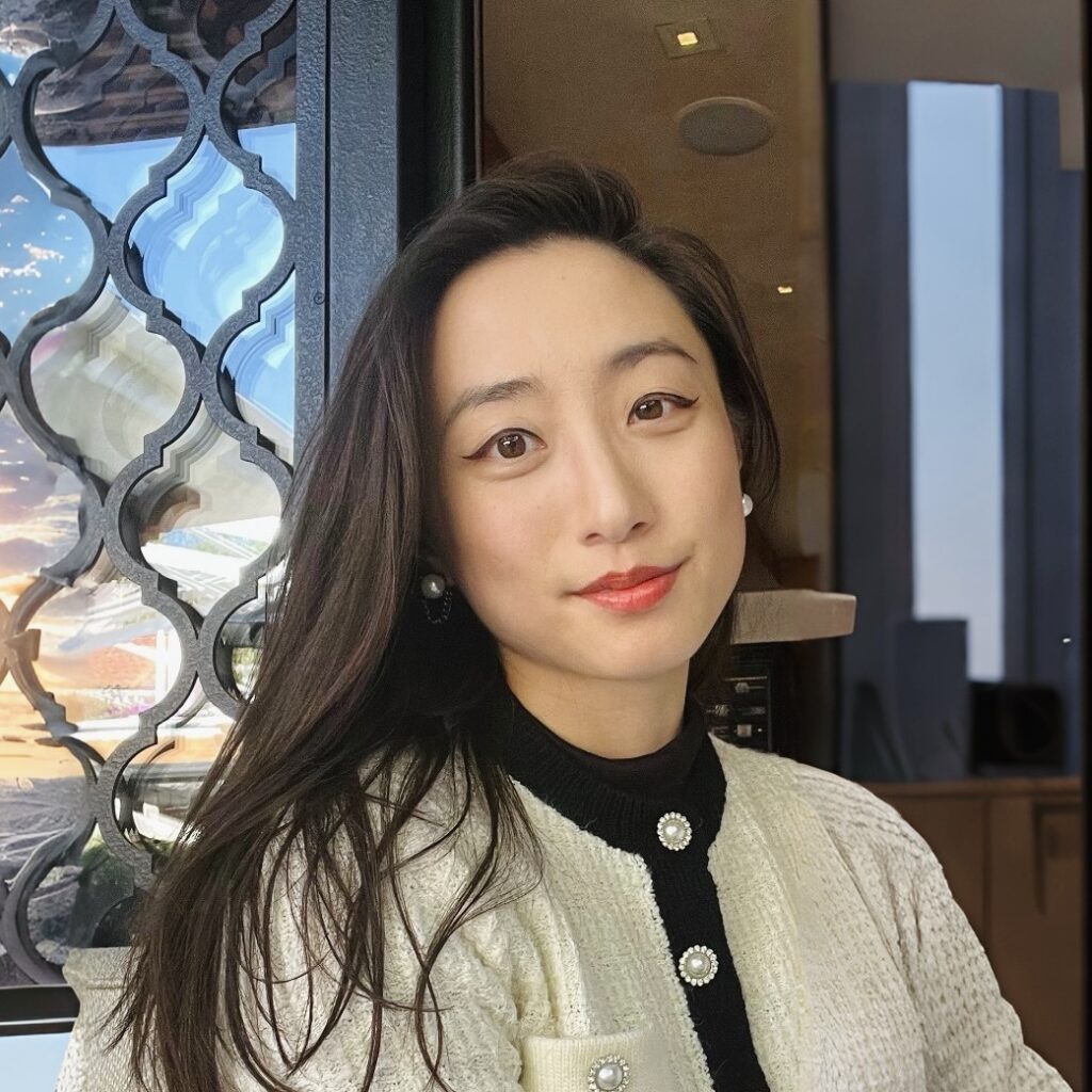 A woman with long dark hair and red lipstick smiles softly at the camera, wearing a cream jacket with pearl buttons and a black turtleneck, seated indoors near a window with patterned glass.