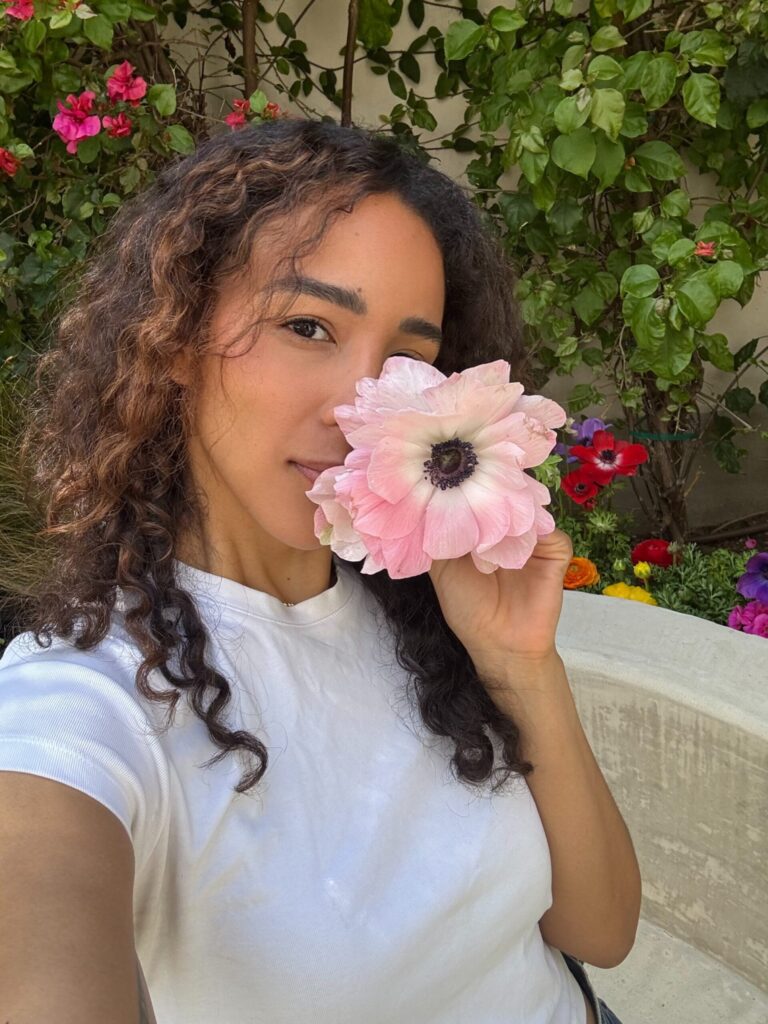 A woman with curly hair wearing a white t-shirt holds a large pink flower in front of her face while taking a selfie in a garden filled with greenery and colorful flowers.