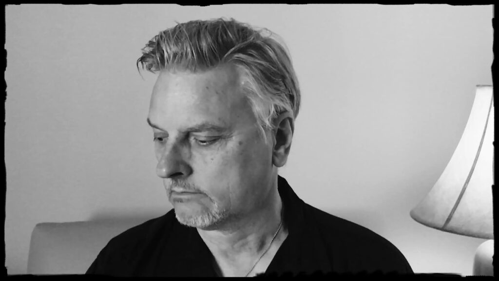 Black and white photo of a man with light hair and a short beard, looking down to his left. He is wearing a dark shirt. A lamp is visible on the right side against a plain wall.