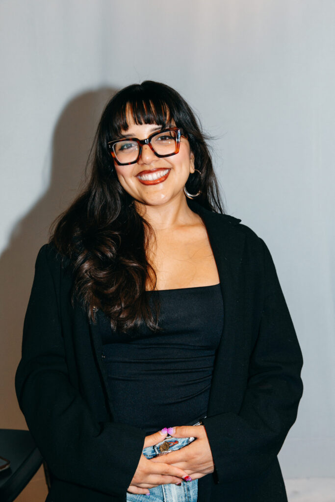 A woman with long dark hair, glasses, and hoop earrings smiles at the camera. She is wearing a black top and black blazer, standing against a plain light background.