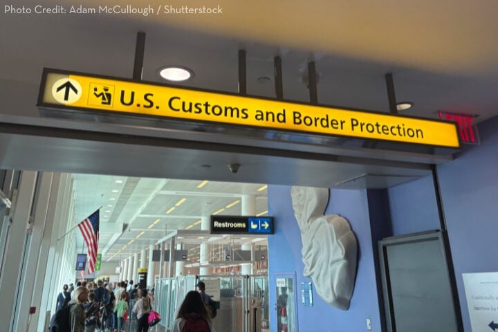A yellow sign in an airport points to U.S. Customs and Border Protection. Travelers walk in the corridor below, and an American flag is visible in the background. A restroom sign also hangs near the customs sign.