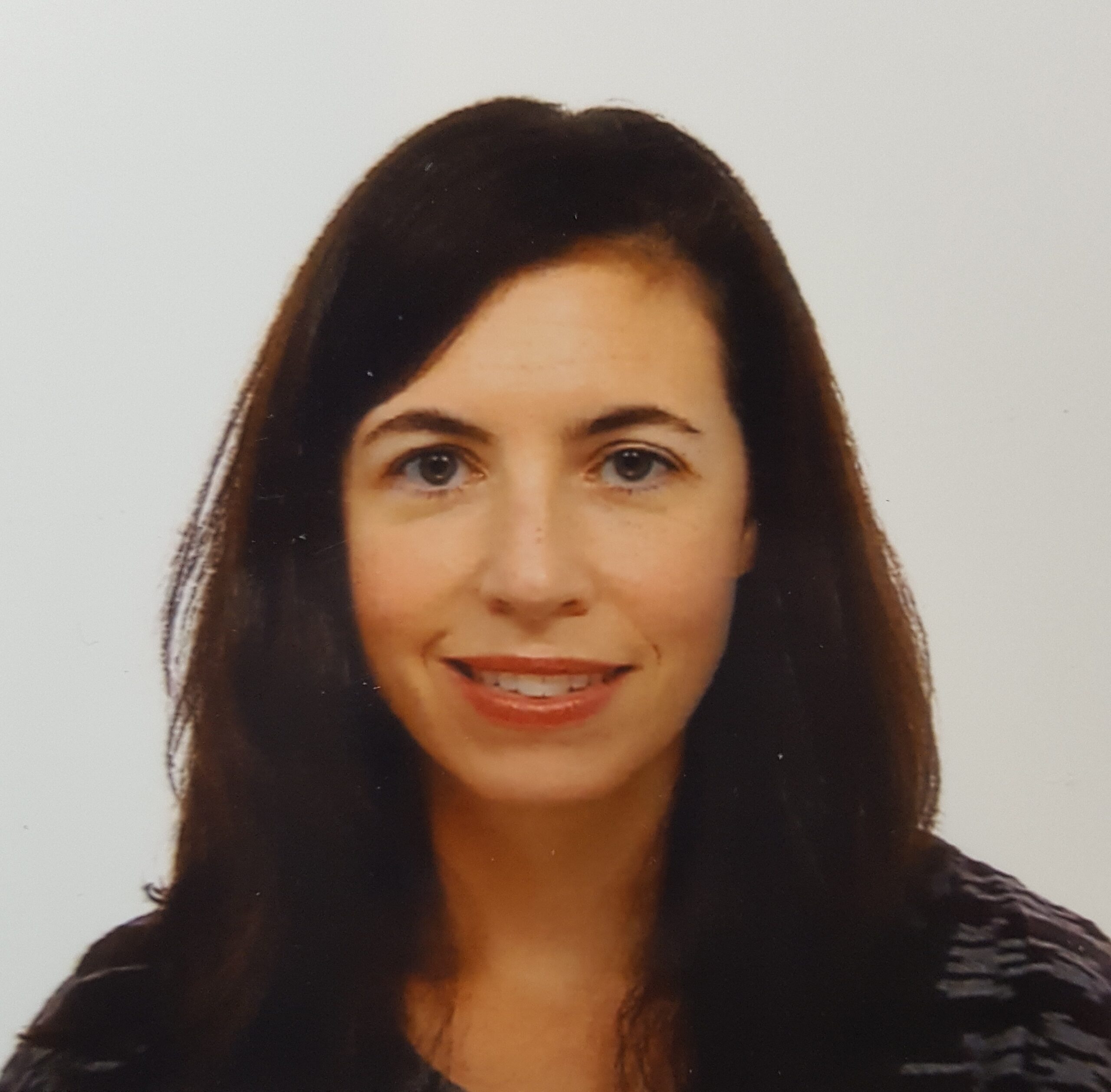 A woman with long dark brown hair, fair skin, and blue eyes is smiling slightly at the camera. She is wearing a dark, patterned top and is photographed against a plain white background.