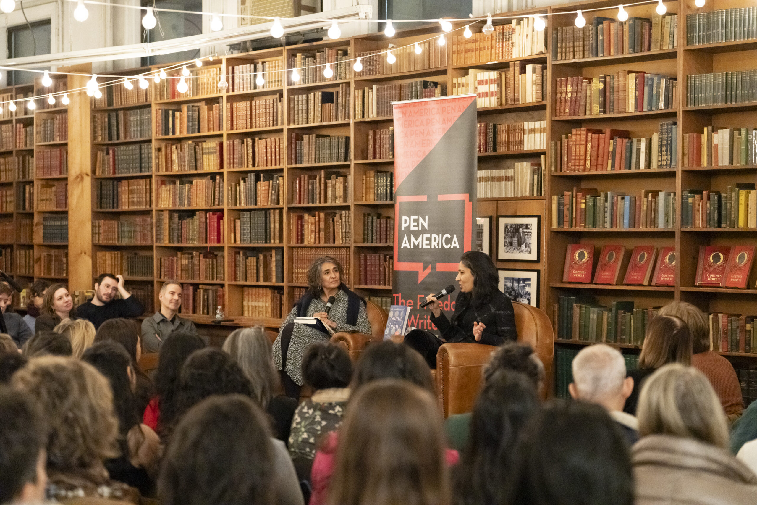 A large audience listens to two women having a conversation onstage in a cozy library with shelves of books, under string lights, at a PEN America event.