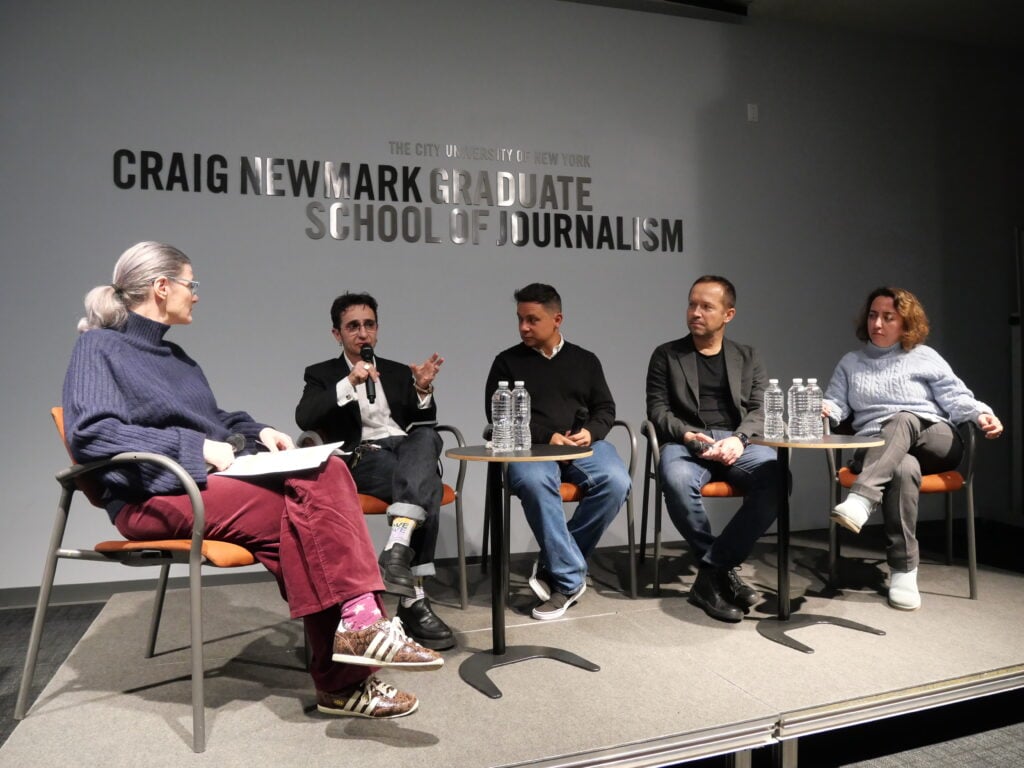 Five people sit on a stage in a panel discussion at the Craig Newmark Graduate School of Journalism. Four listen while one person speaks into a microphone; water bottles are on the tables in front of them.