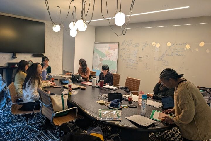 Seven people sit and work around a conference table in a meeting room with laptops, notebooks, and drinks; notes and diagrams are written on a large whiteboard behind them. A large screen and ceiling lights are visible.
