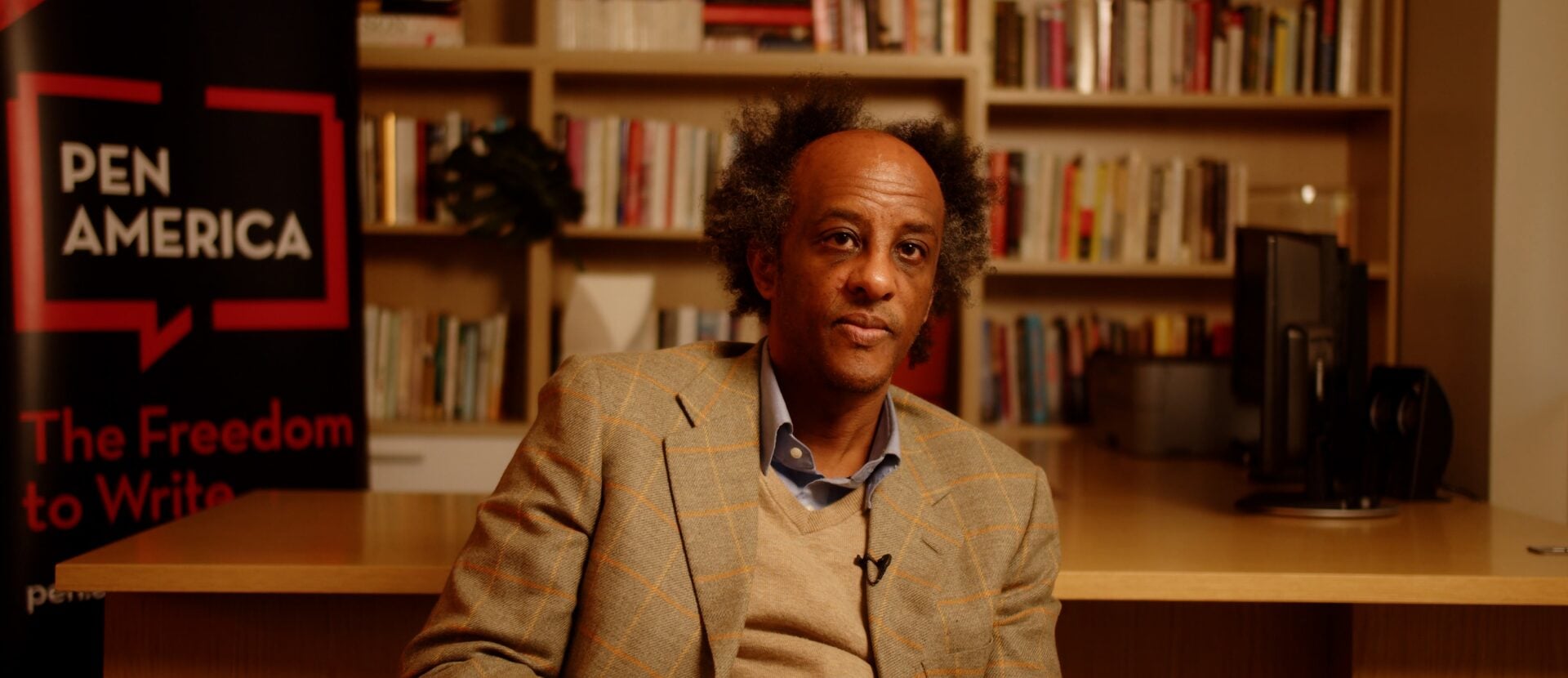A man with graying curly hair sits at a wooden desk in front of bookshelves, wearing a tan plaid blazer and blue shirt. A PEN America sign with the words The Freedom to Write is visible in the background.