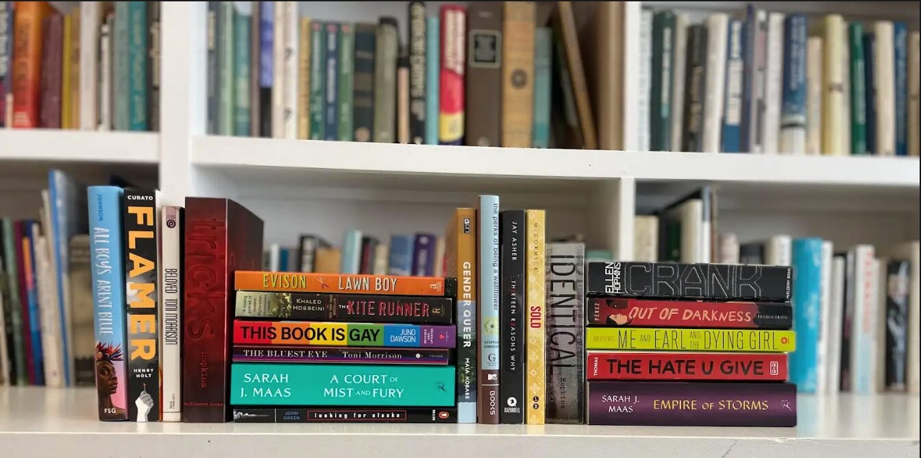 A stack of 20 banned books, most of them with LGBTQ+ and BIPOC representation, including titles like Genderqueer by Maia Kobabe and The Hate U Give by Angie Thomas.