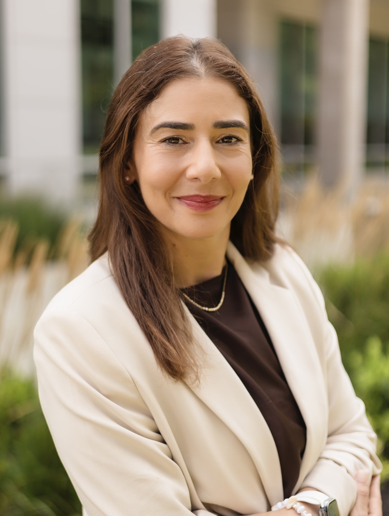 A woman with long brown hair wearing a cream blazer and brown top smiles with arms crossed, standing outdoors in front of a blurred building and greenery.