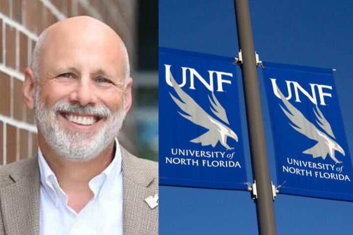 A smiling bald man with a gray beard stands by a brick wall; next to him are two blue banners with a white osprey logo and text reading “UNF University of North Florida” against a clear blue sky.