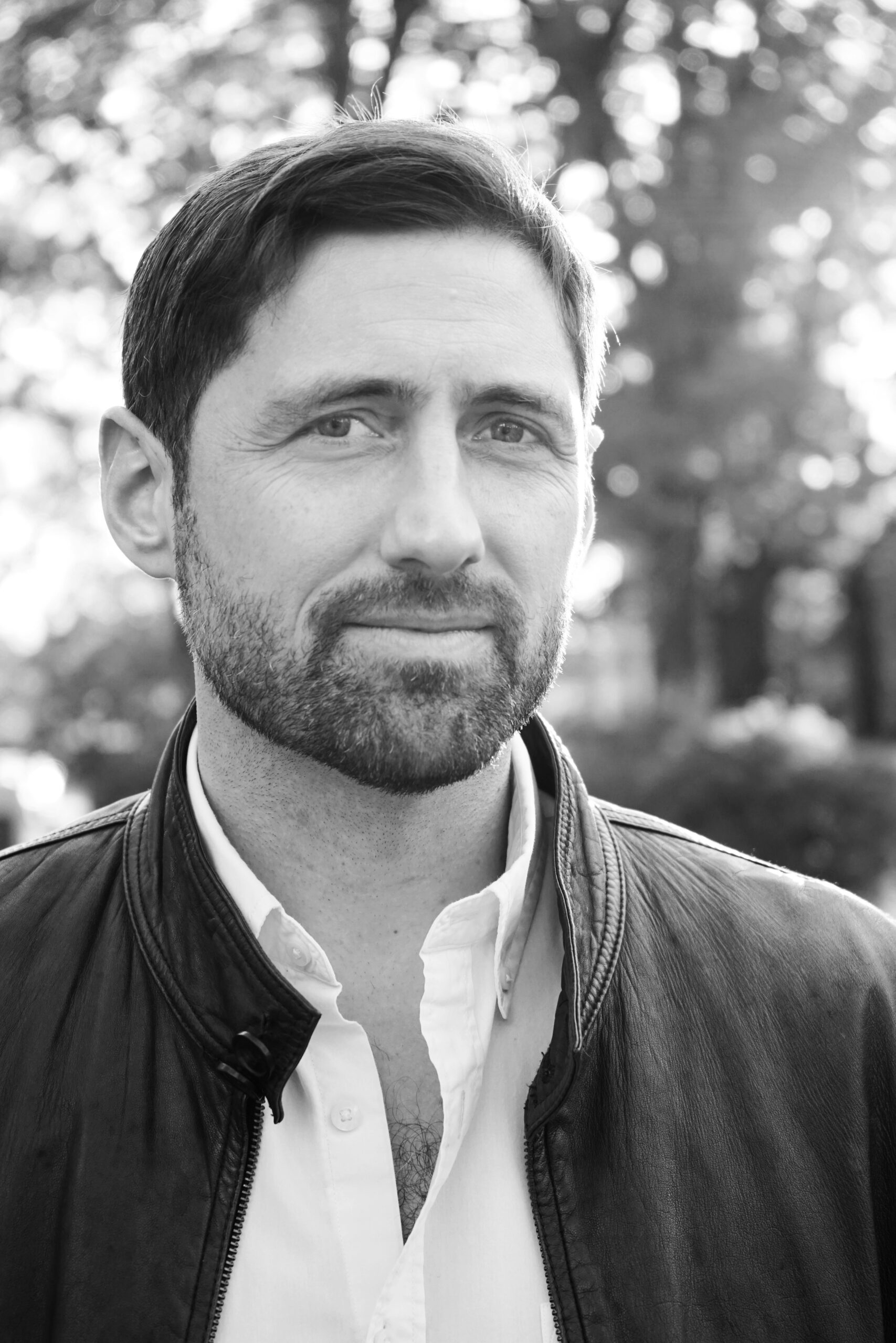 A man with short dark hair and a beard wears a leather jacket over a white shirt, standing outdoors with trees and sunlight in the blurred background.