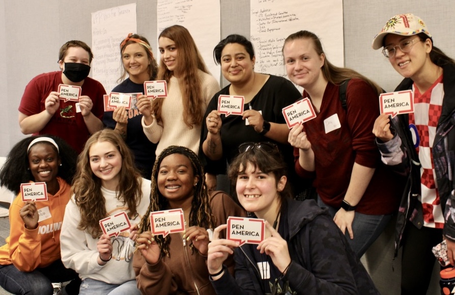 A group of ten smiling people pose indoors, holding up red and white PEN America stickers. Some are kneeling in front, others stand behind. Sheets of paper with writing hang on the wall behind them.