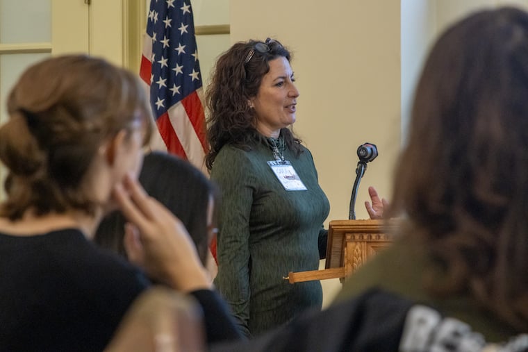 A woman speaks at a podium in front of an American flag, addressing an audience. She wears a name tag and gestures with her hand, while listeners sit in the foreground.