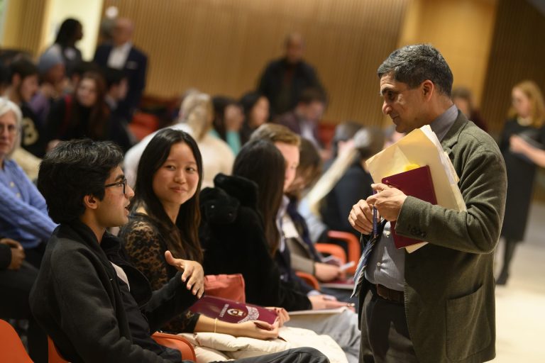 A man holding folders talks to a seated young man in a crowded auditorium. Other people sit and chat around them, some looking forward and others engaging in conversation. The atmosphere is lively and informal.