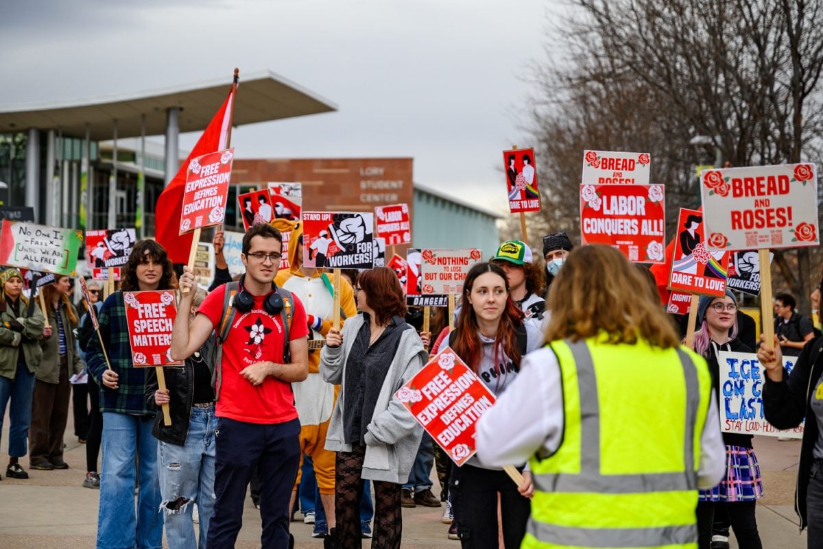 A group of people hold signs reading “Free Speech Matters,” “Labor Conquers All,” and “Bread and Roses!” at an outdoor protest. One person in a yellow vest faces the crowd. A library building is visible in the background.