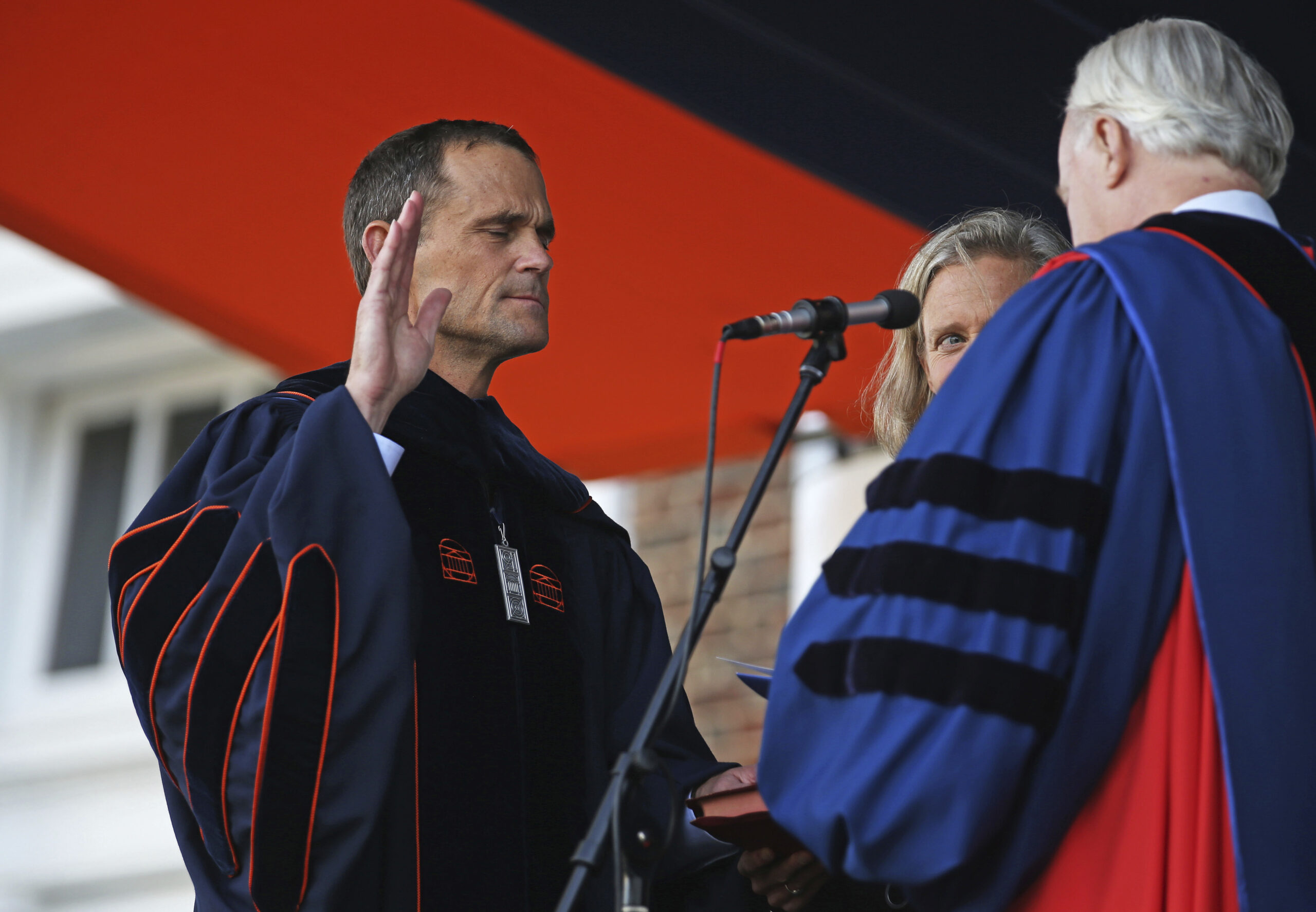 A man in academic regalia raises his right hand while being sworn in on stage during a formal ceremony, with two other people in academic gowns standing beside him.