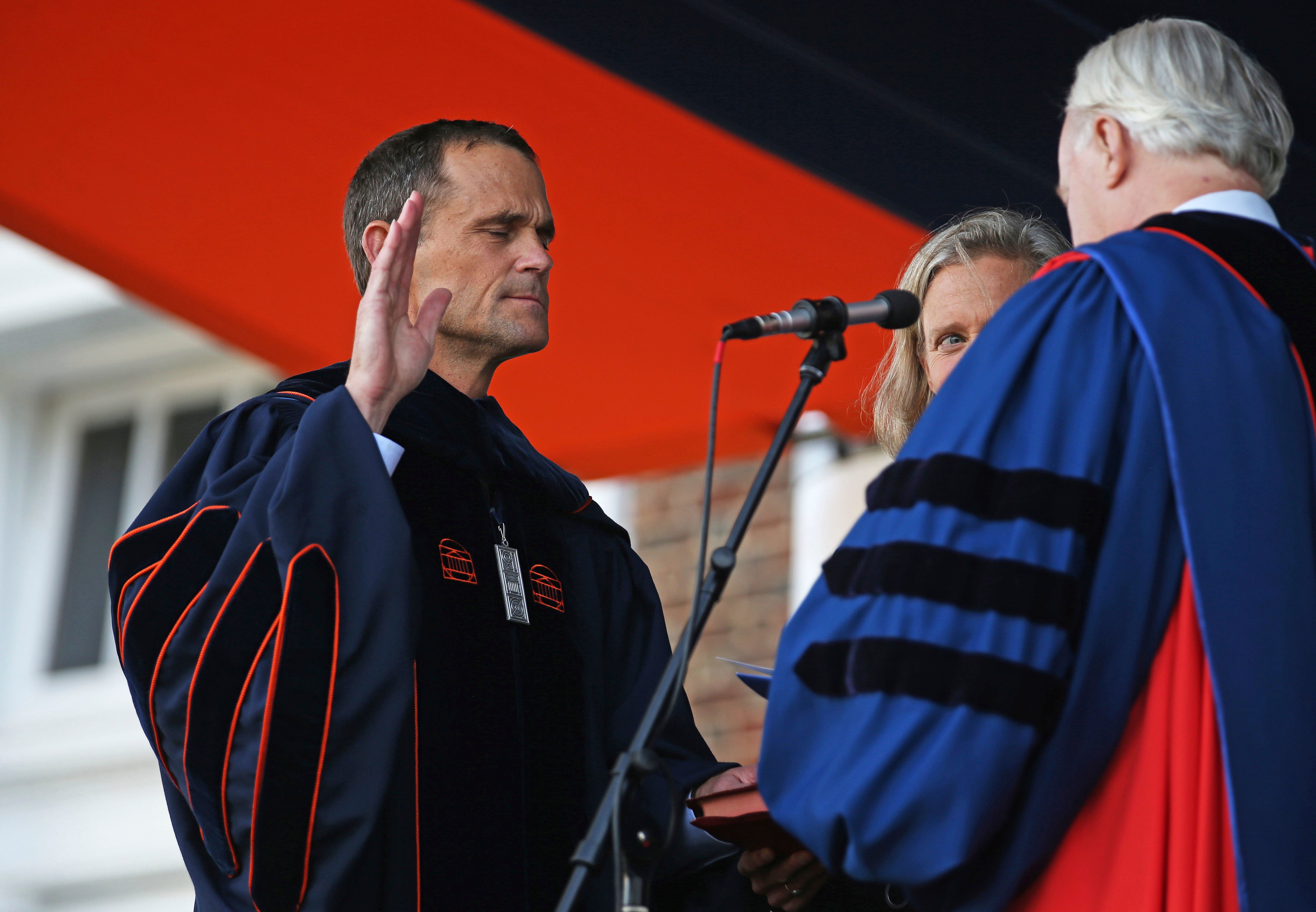 A man in academic regalia raises his right hand while being sworn in on stage during a formal ceremony, with two other people in academic gowns standing beside him.
