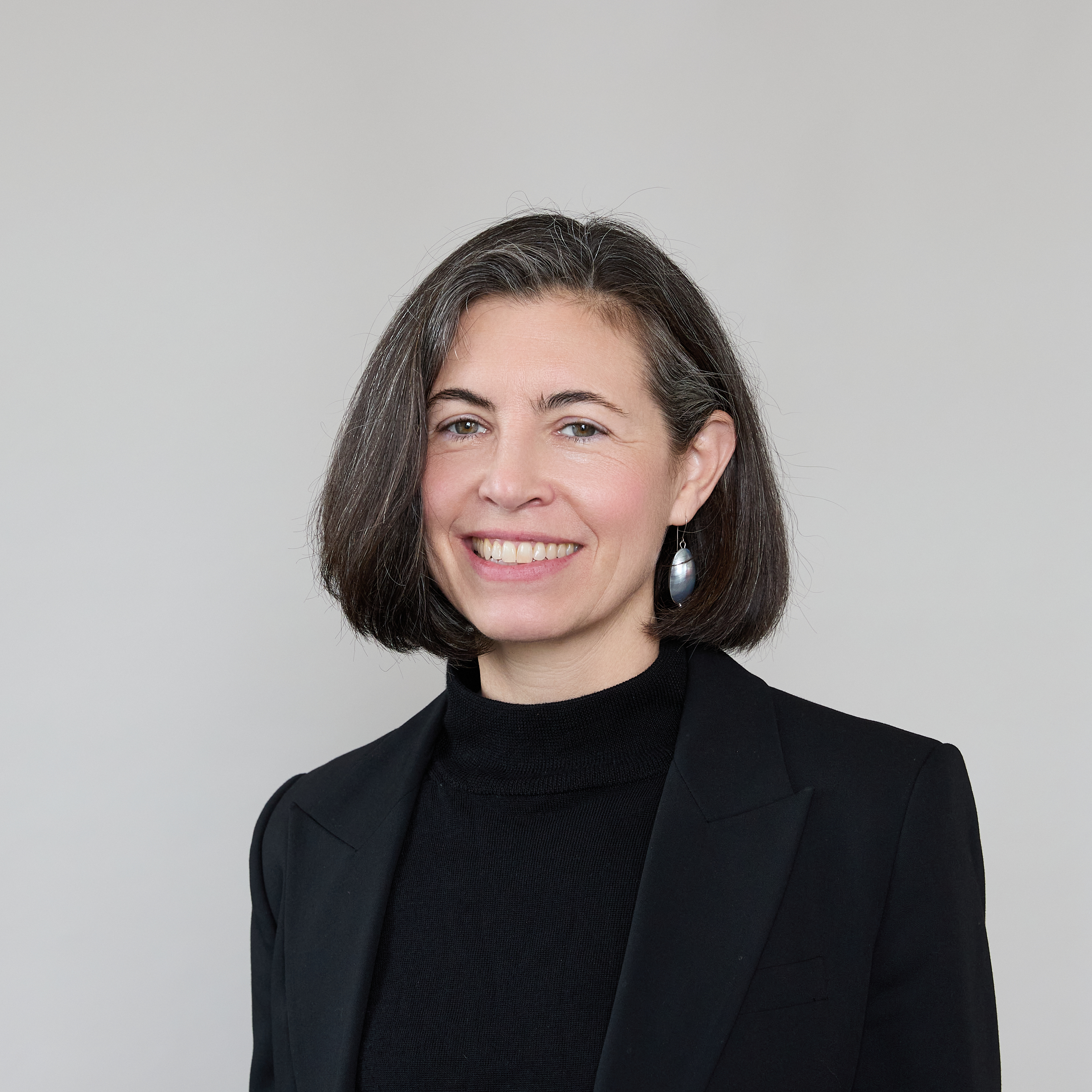 A woman with straight shoulder-length dark hair, wearing a black blazer and black top, smiles at the camera against a plain light grey background.