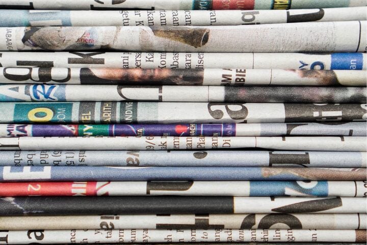 A close-up of stacked, folded newspapers with visible text, headlines, and colored images creating a layered, textured pattern. The newspapers overlap horizontally and display various fragments of print.