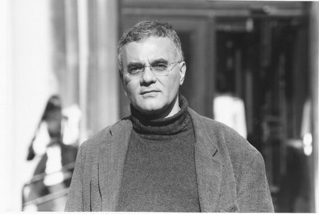 A man with short gray hair and glasses, wearing a turtleneck sweater and a corduroy jacket, stands outdoors in front of a blurred building entrance, looking directly at the camera.