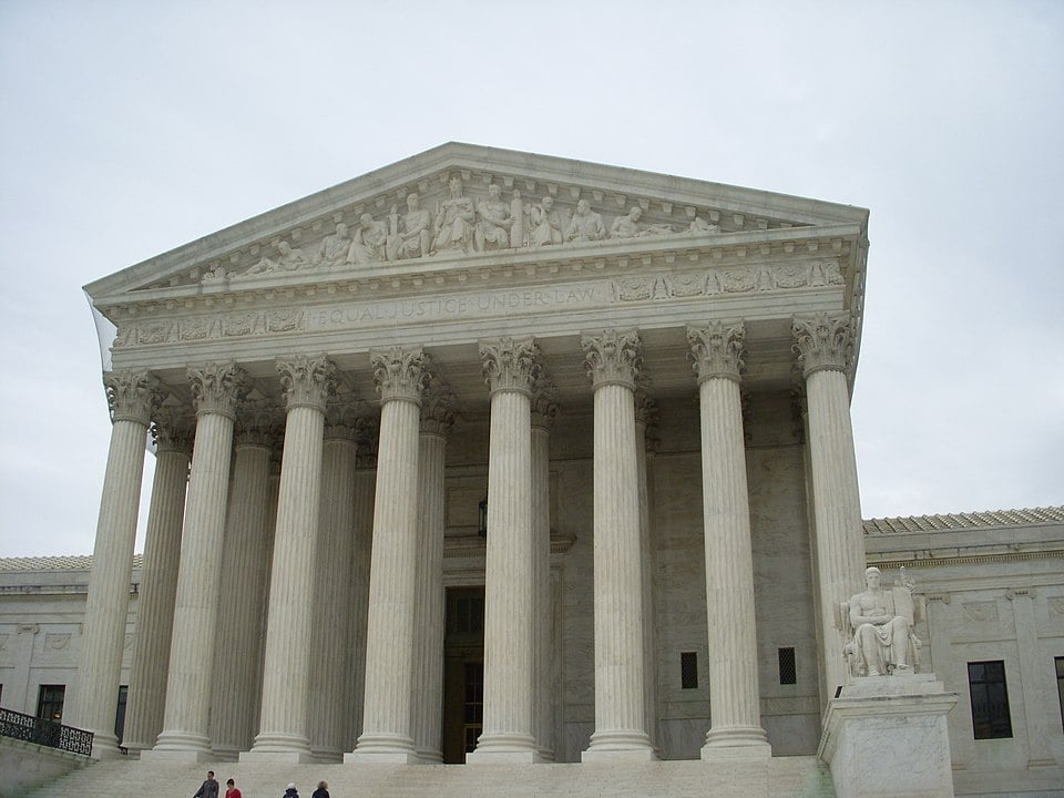 The image shows the front of the United States Supreme Court building in Washington, D.C., featuring large columns, detailed carvings on the pediment, and a statue on the right side of the steps under a cloudy sky.