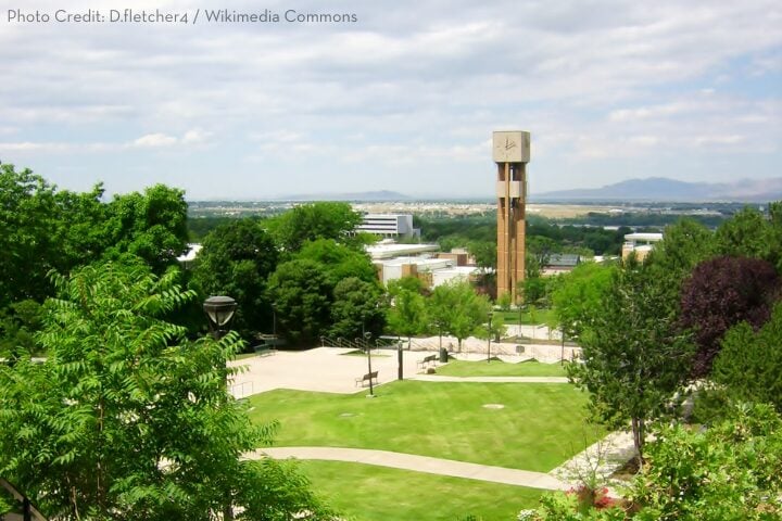 View of a green campus with trees, walkways, open lawn, and a tall clock tower in the center. Buildings and distant mountains are visible under a partly cloudy sky.