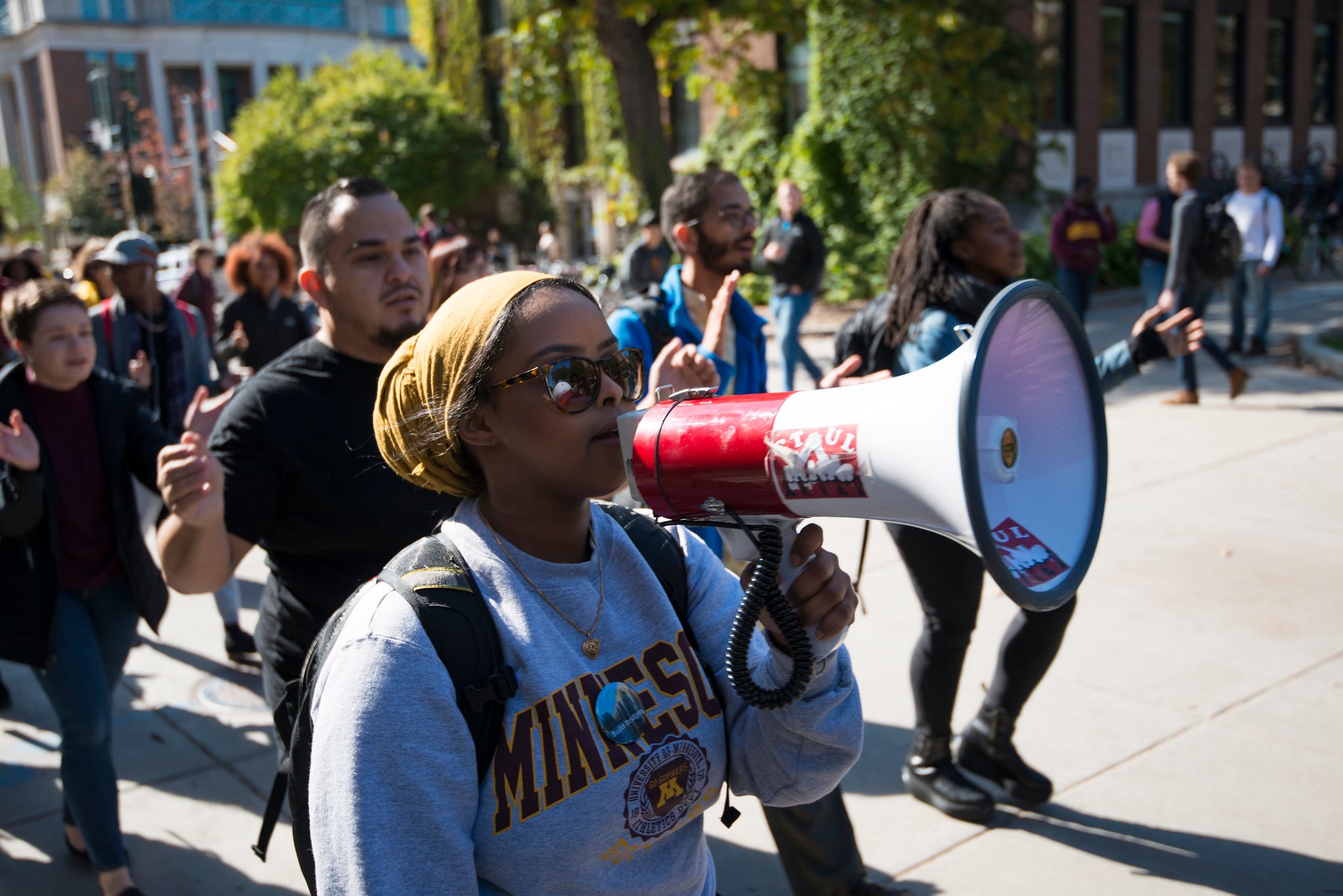 A woman wearing sunglasses and a Minnesota sweatshirt leads a group of people in a protest, speaking into a red and white megaphone as they march outdoors on a sunny day.