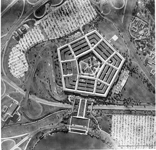 Aerial black-and-white photo of the Pentagon building, featuring its distinct five-sided shape, surrounded by roads, parking lots filled with cars, and green areas.