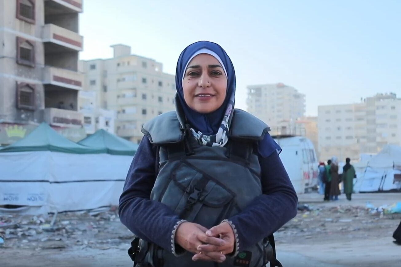 A woman wearing a hijab and protective vest stands smiling in front of tents and damaged buildings in an urban area, with people walking in the background.